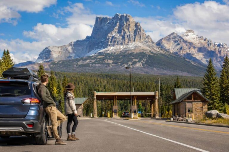 Couple leaning against a blue SUV at a mountain park entrance with snowy peaks in the background.