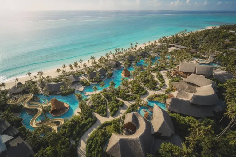 Aerial view of a tropical resort with thatched-roof villas, winding water slides, and turquoise ocean beyond a palm-lined shore.