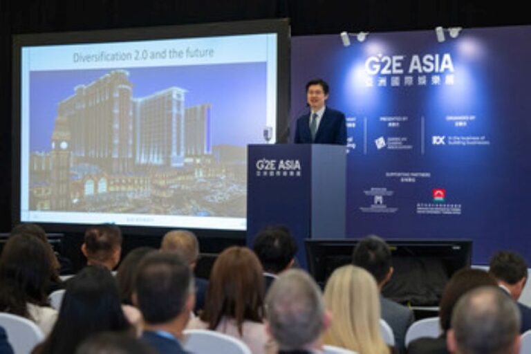 Presenter in a suit at a podium giving a conference talk, slide titled 'Diversity 2.0 and the future' seen on a large screen