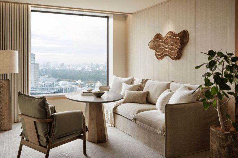 Cozy neutral living room with a beige sofa, round wooden table, armchair, potted plant, and large window overlooking the city skyline.