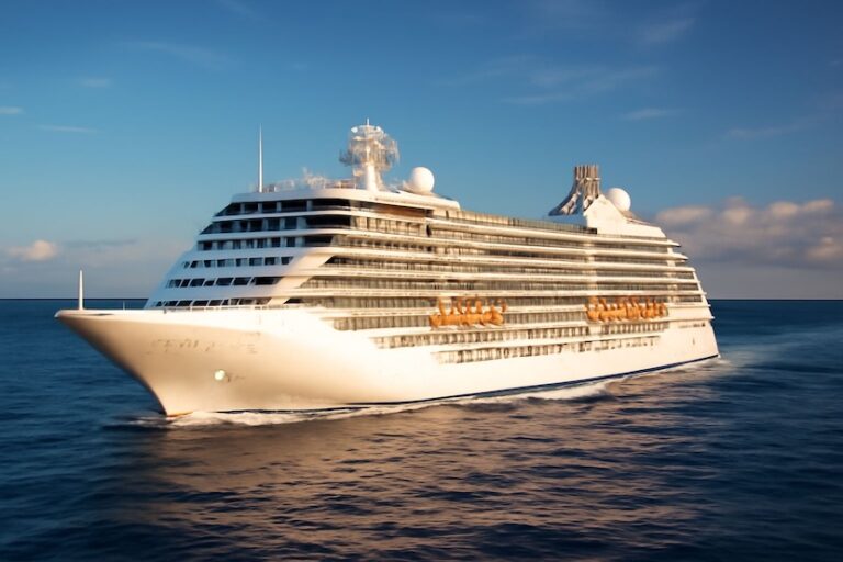 Large white cruise ship sailing on calm blue ocean under a clear sky, with multiple decks and orange lifeboats along the side.