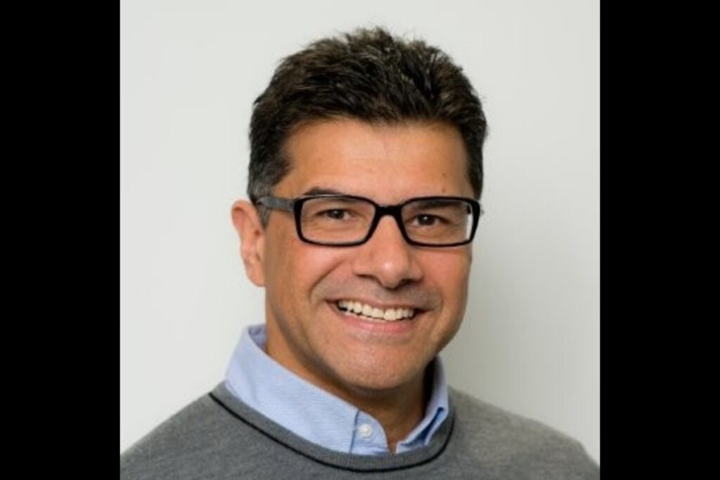 Smiling man with black glasses wearing a gray sweater over a light blue collared shirt, facing camera.