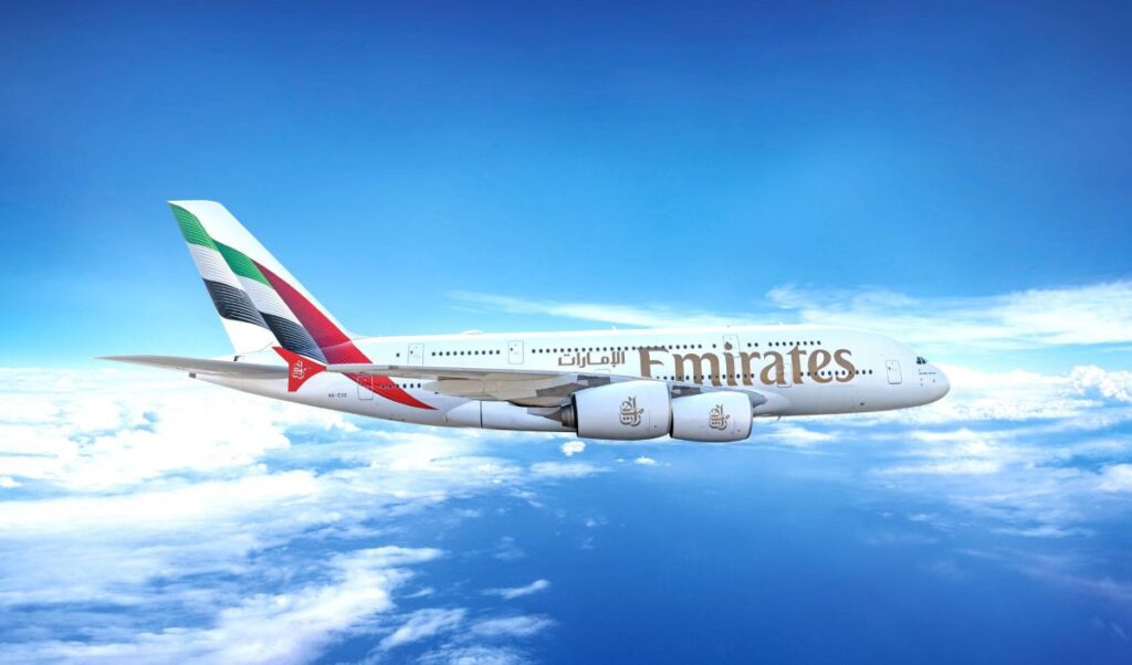 Emirates passenger airplane in midflight above white clouds and a blue sky, tail featuring multi-color design of the UAE flag.