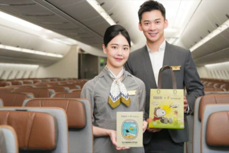 Two flight attendants stand in an airplane cabin, holding a safety card and a Snoopy-themed gift box.