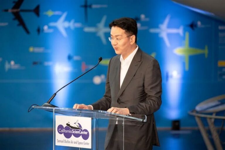 Man in a dark suit delivers a speech at a clear podium with a California Science Center logo, microphone present, blue backdrop with airplane silhouettes
