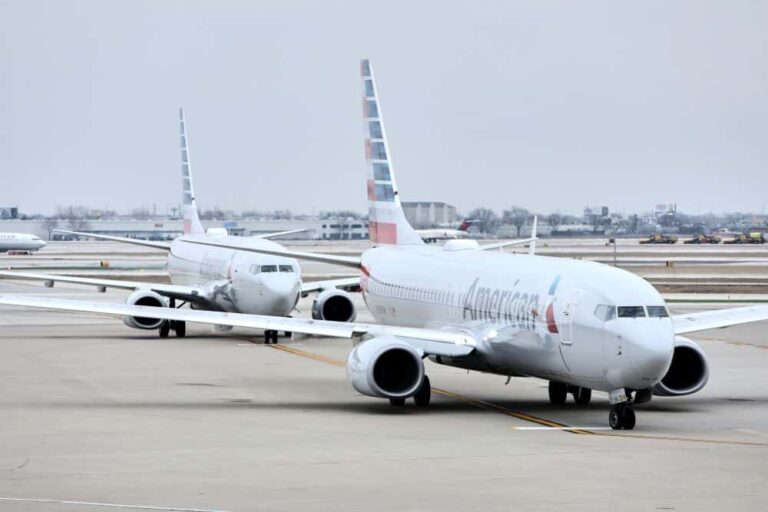 Two American Airlines jets taxi on the runway at an airport, with distant terminal buildings visible in a pale sky.