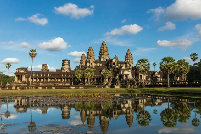 Angkor Wat temple complex with central towers, palm trees, and a moat reflecting the sky and structures.