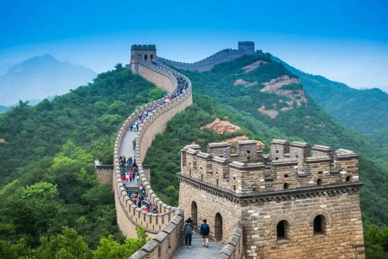 Tourists walk along a winding stone path of the Great Wall across green hills and watchtowers in the distance.