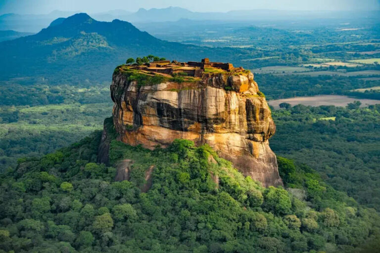Rock fortress perched on a towering sandstone cliff, with ruins on the sunlit summit and dense forest below.