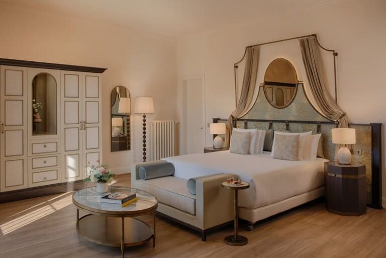 A luxurious hotel room with a king bed under an arched, draped headboard, beige and cream decor, and matching nightstands and lamps on either side.