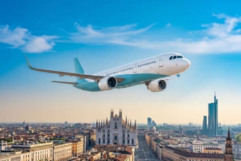 White passenger jet flies over a city with a prominent gothic cathedral and modern skyline beneath a blue sky.