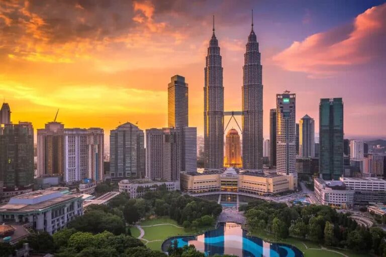Sunset over Kuala Lumpur with the Petronas Twin Towers and city skyline, a green park and water feature in the foreground.