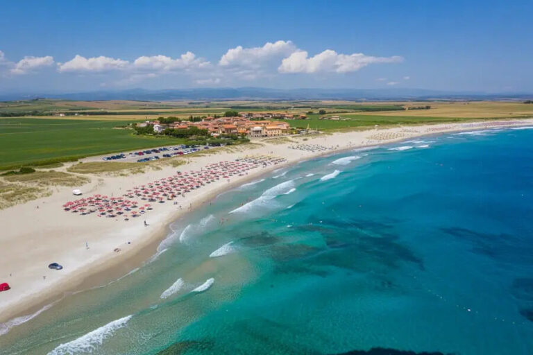 Aerial view of a sandy beach with rows of red umbrellas, turquoise water, and a small village inland. (informative)