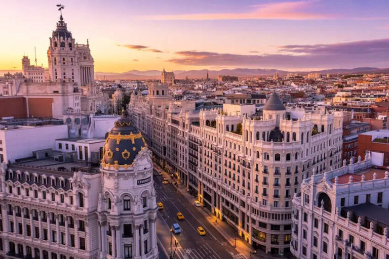 Madrid city skyline at sunset with ornate historic buildings and a busy street below, golden light across the horizon.