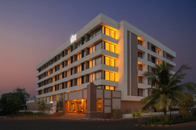 Modern multi-story hotel with warmly lit windows at dusk, palm trees in front.