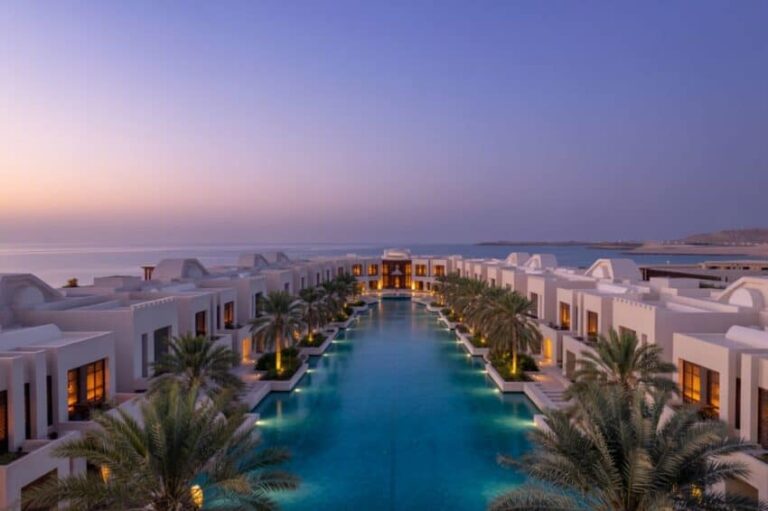 Resort pool lined with palm trees between white villas at sunset, calm sea in the background.