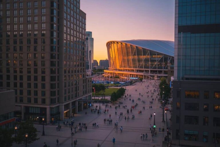 Sunset-lit city plaza with a curved, glass-fronted stadium glowing orange in the background and crowds walking below.