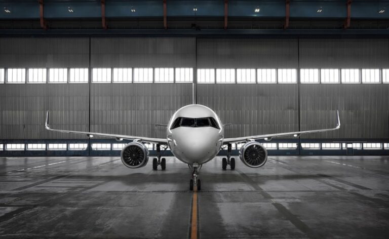 Front view of a commercial airplane parked inside a hangar, facing the camera with engines on each wing and a yellow centerline on the tarmac