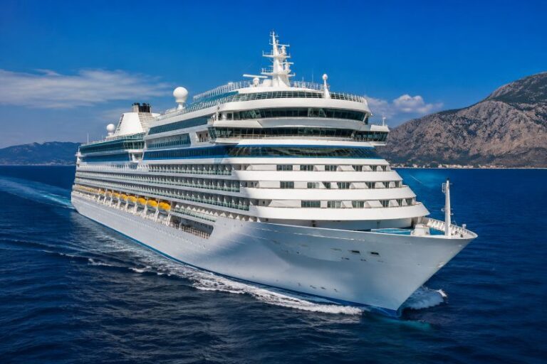 White cruise ship with multiple decks sailing in deep blue sea, mountains in the background under a clear sky.