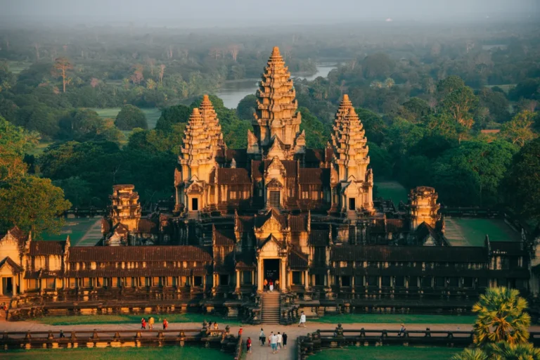 Angkor Wat temple complex in Cambodia with five central towers rising above a long facade, visitors walking along the front path beside the moat.