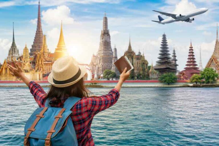 Traveler with a blue backpack and straw hat raises arms toward a temple-lined skyline across a river, holding a passport; airplane flies overhead.