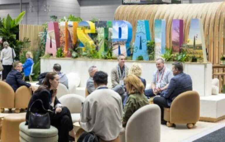 Group of people in a lounge area at a conference, chatting in front of a tall, colorful sign behind them in an expo hall.