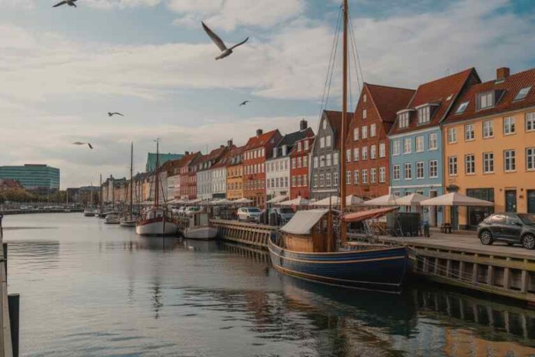 Colorful waterfront town with boats docked along a canal and a row of bright houses lining the quay, under a cloudy sky.