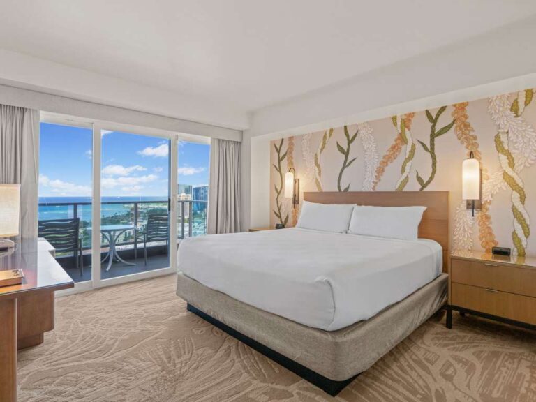 Hotel room with a king bed, floral wallpaper, and a balcony overlooking the ocean. (informative)