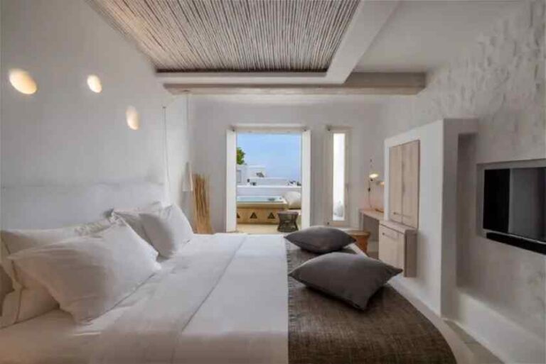 Bright white hotel bedroom with a large bed, gray floor cushions, and a balcony door revealing a blue sea view outside.