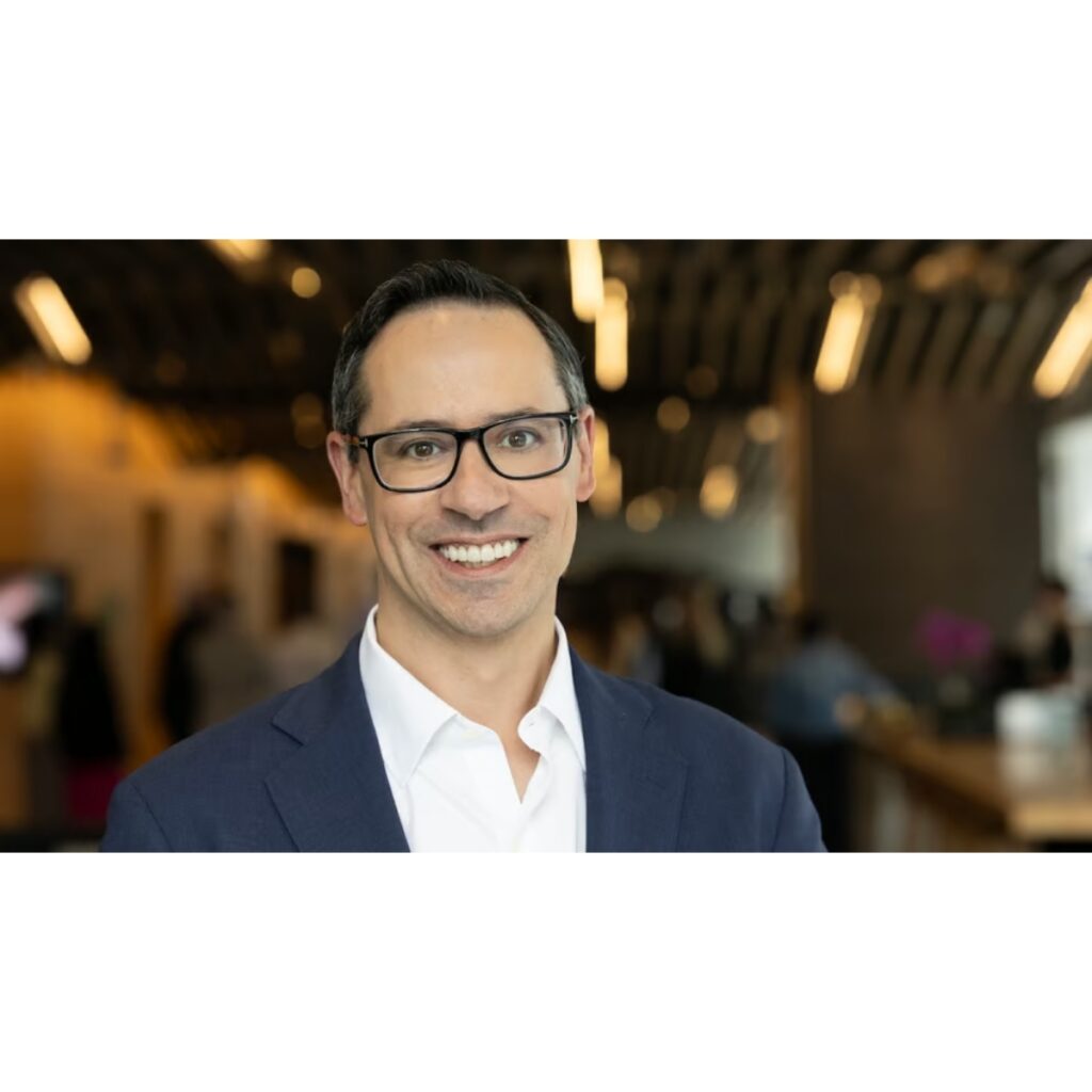 Professional headshot: smiling man in a navy suit and glasses in a modern lobby setting.