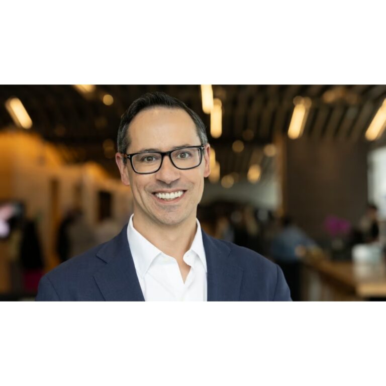 Professional headshot: smiling man in a navy suit and glasses in a modern lobby setting.