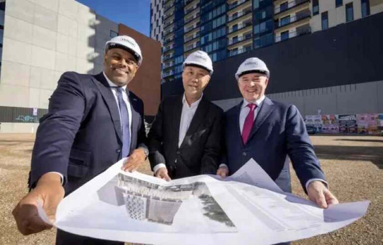Three businessmen in suits and hard hats examine large blueprints at a construction site with a high-rise building behind them.