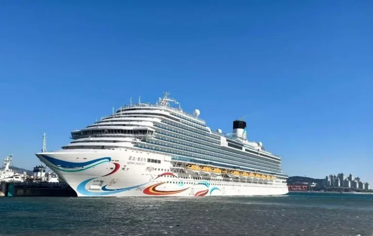 Large white cruise ship docked at a port under a clear blue sky, with colorful wave graphics along the hull.