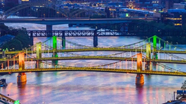 Colorful night view of multiple illuminated bridges spanning a river, with green, yellow, and red lights reflecting on the water.