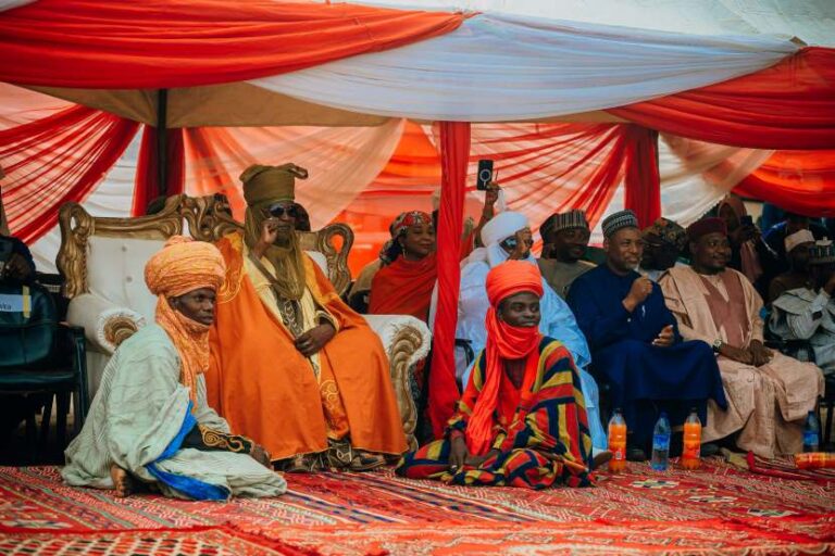 Dignitaries in vibrant traditional outfits sit under a red-and-white ceremonial canopy, part of a cultural gathering or wedding.