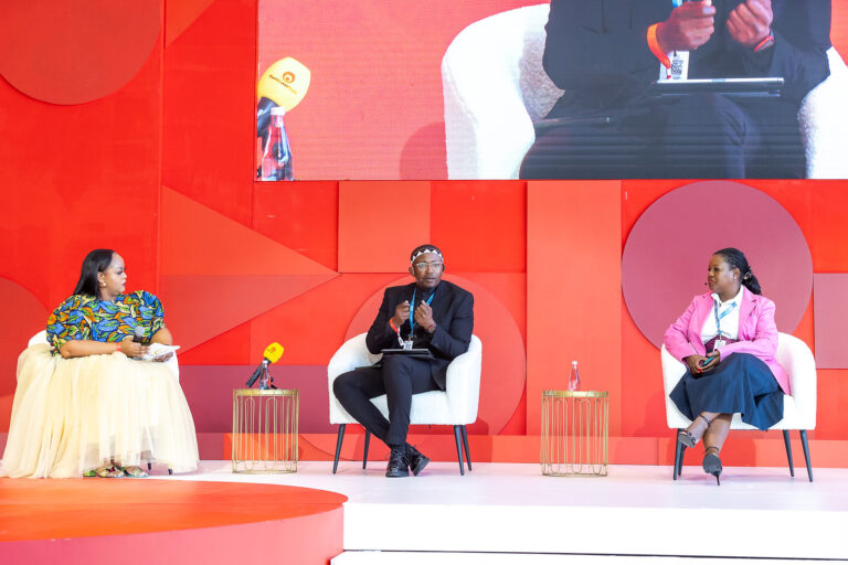 Panel discussion with three speakers on a red stage; left woman in colorful dress holds a microphone, center man in a black suit speaks, right woman in a pink blazer listens.