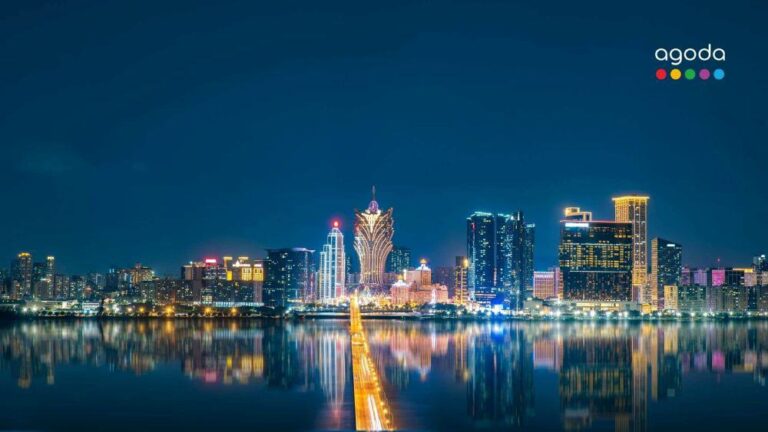Night city skyline with a brightly lit bridge spanning calm water; reflections glow in the river, Agoda logo in the top-right corner