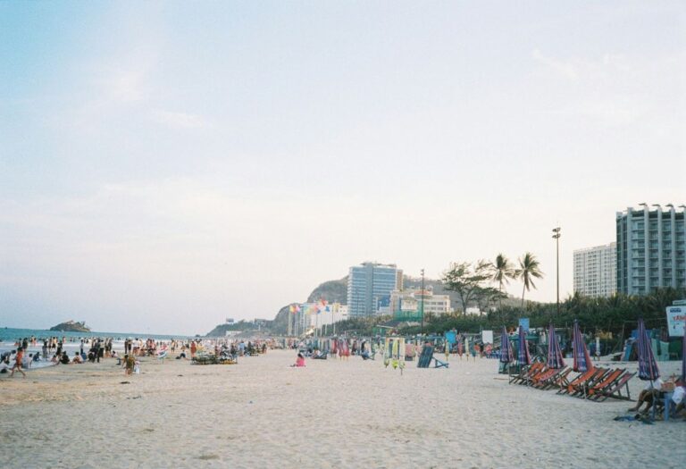 Busy beach with families and sun loungers under colorful umbrellas, high-rise hotels along the coast in the background.