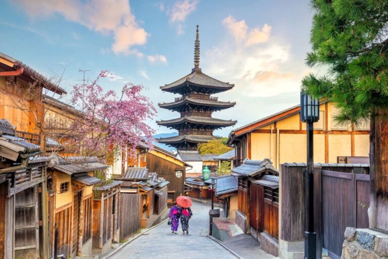 Traditional Japanese street with wooden houses and a five-story pagoda in the distance; two people walk under a red umbrella.