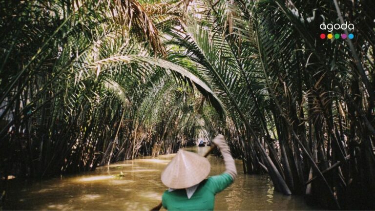 Person in a green shirt paddles a small boat through a narrow, shaded waterway lined with tall palm trees; agoda logo in the corner.