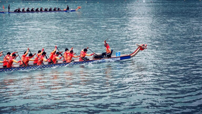 Dragon boat team in red shirts paddling in unison on blue water, dragon-head boat with drummer at front and paddlers in a race.