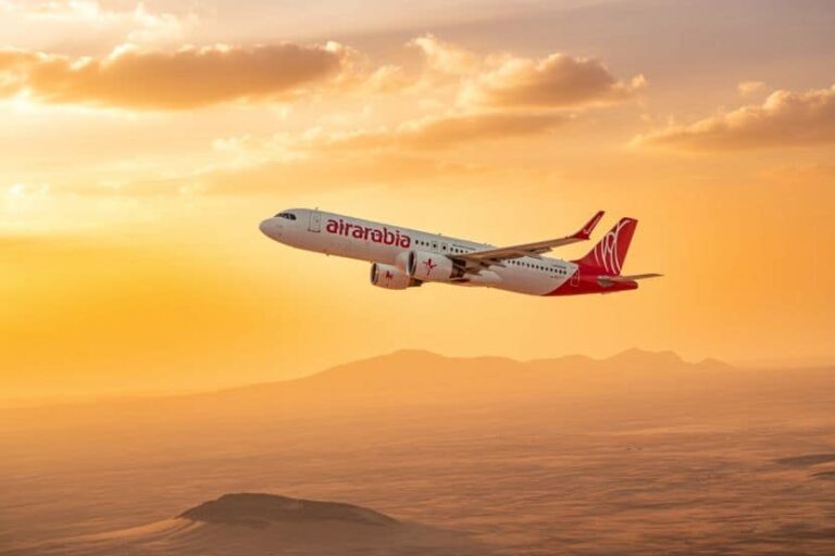 Air Arabia passenger jet flying over a desert landscape at sunset with orange sky and mountains in the distance, silhouetted dunes below.