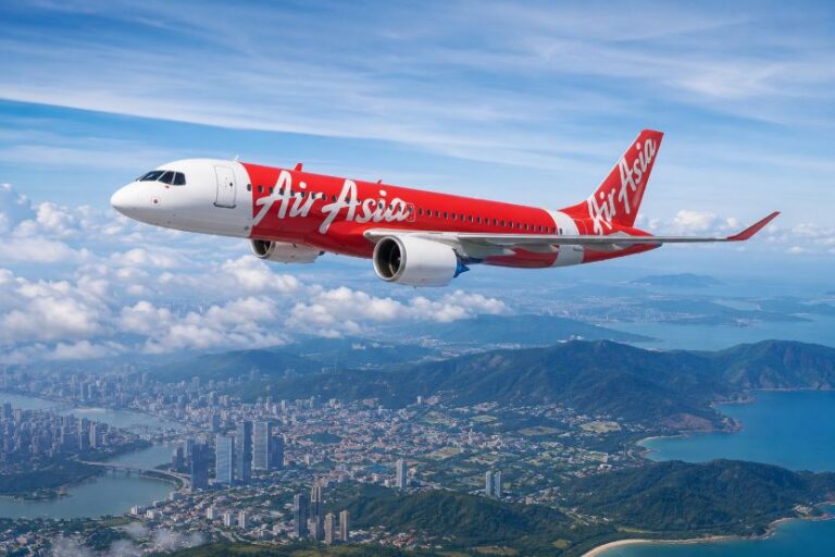Red AirAsia passenger jet in flight above a coastal city and hills, with a bright blue sky and scattered clouds.