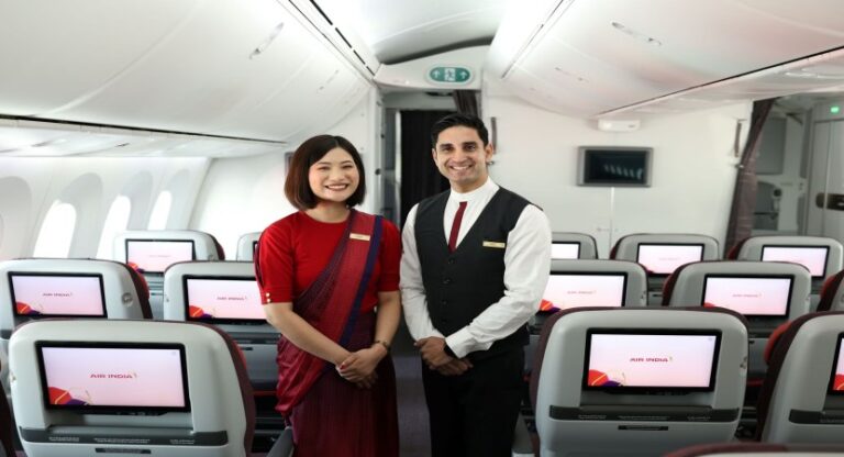 Two Air India flight attendants stand in an airplane cabin, smiling at the camera in matching uniforms.