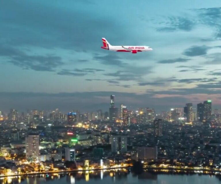 Air India passenger jet flying over a nighttime city skyline with tall buildings and a river reflecting lights.