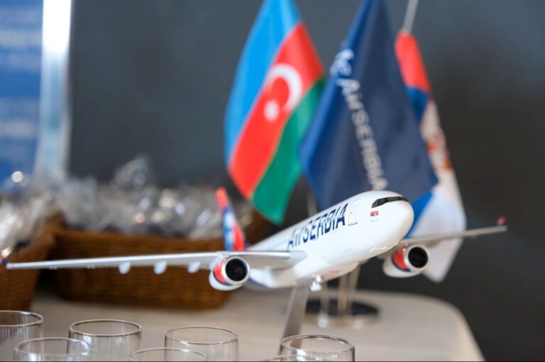 Model airplane on display with blue, red, and green flags in the background; foreground shows drinking glasses on a table edge.