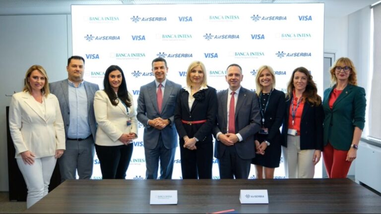 Ten professionals in business attire pose for a group photo at a corporate event in front of a branded backdrop with logos.