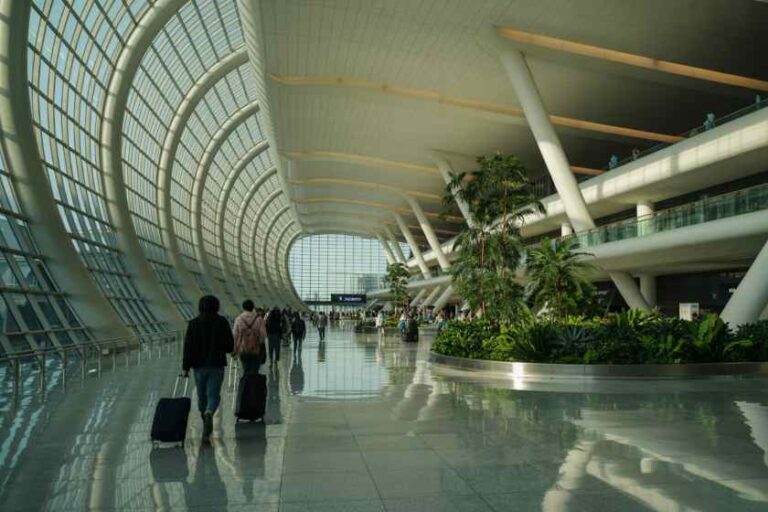 Passengers with luggage walk through a bright, curved glass airport terminal with indoor plants.