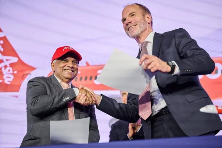 Two businessmen shake hands on stage after signing documents, with a red airplane backdrop behind them.