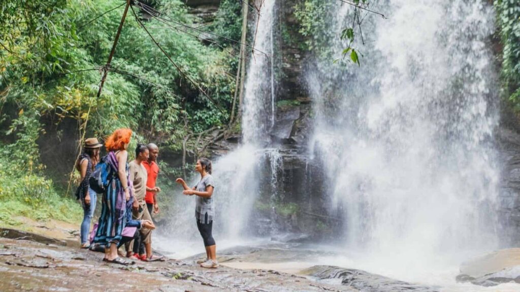 Group of travelers listening to a guide near a cascading waterfall in a lush jungle setting.